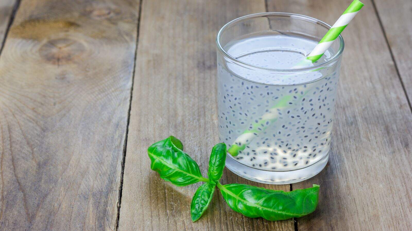 A glass of basil seed water with a green and white striped straw, placed on a wooden surface. Two fresh basil leaves are positioned next to the glass. The drink contains visible basil seeds suspended in the liquid.