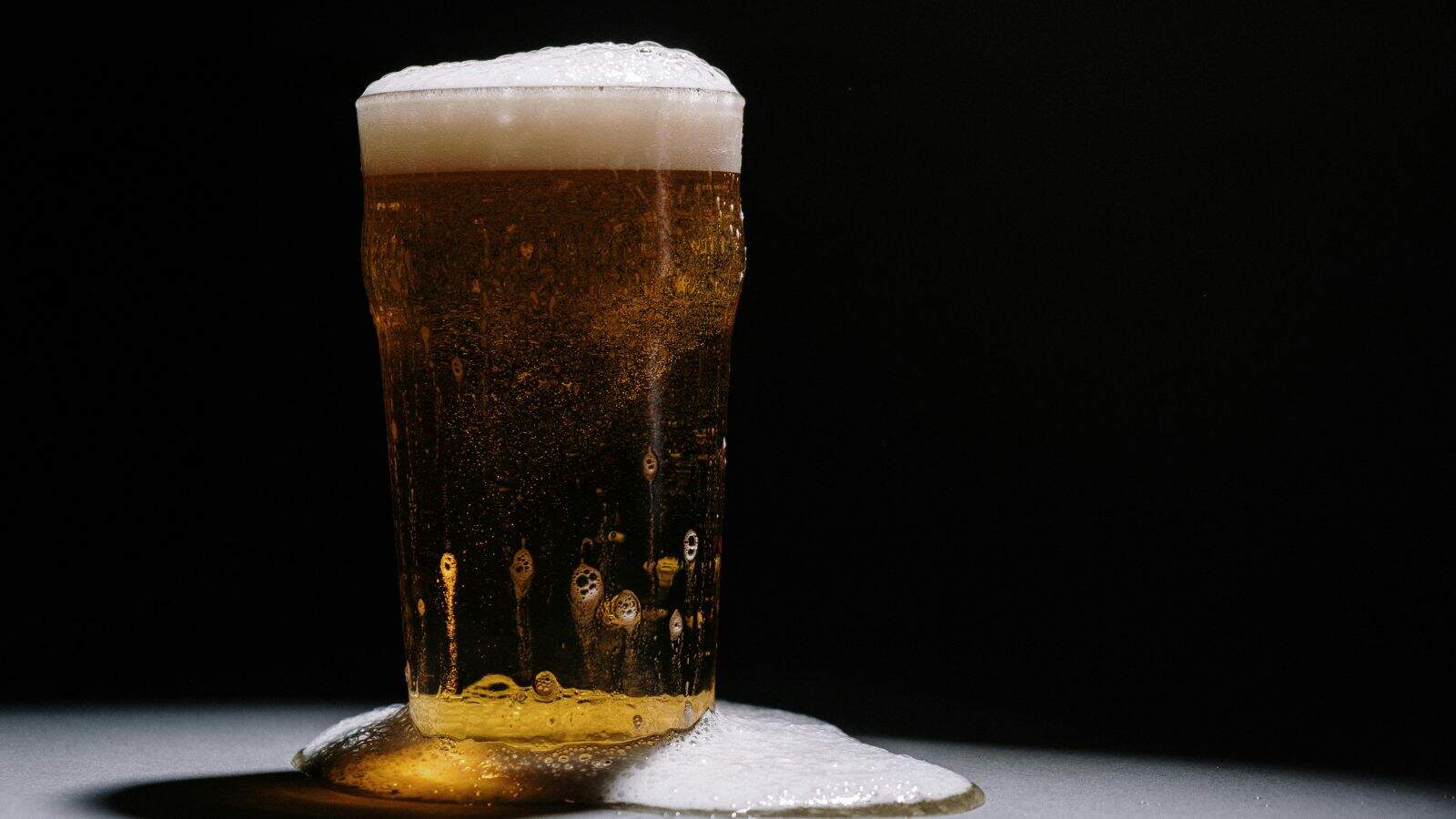 A frothy beer in a glass sits on a surface, with foam spilling over the sides, set against a dark background.