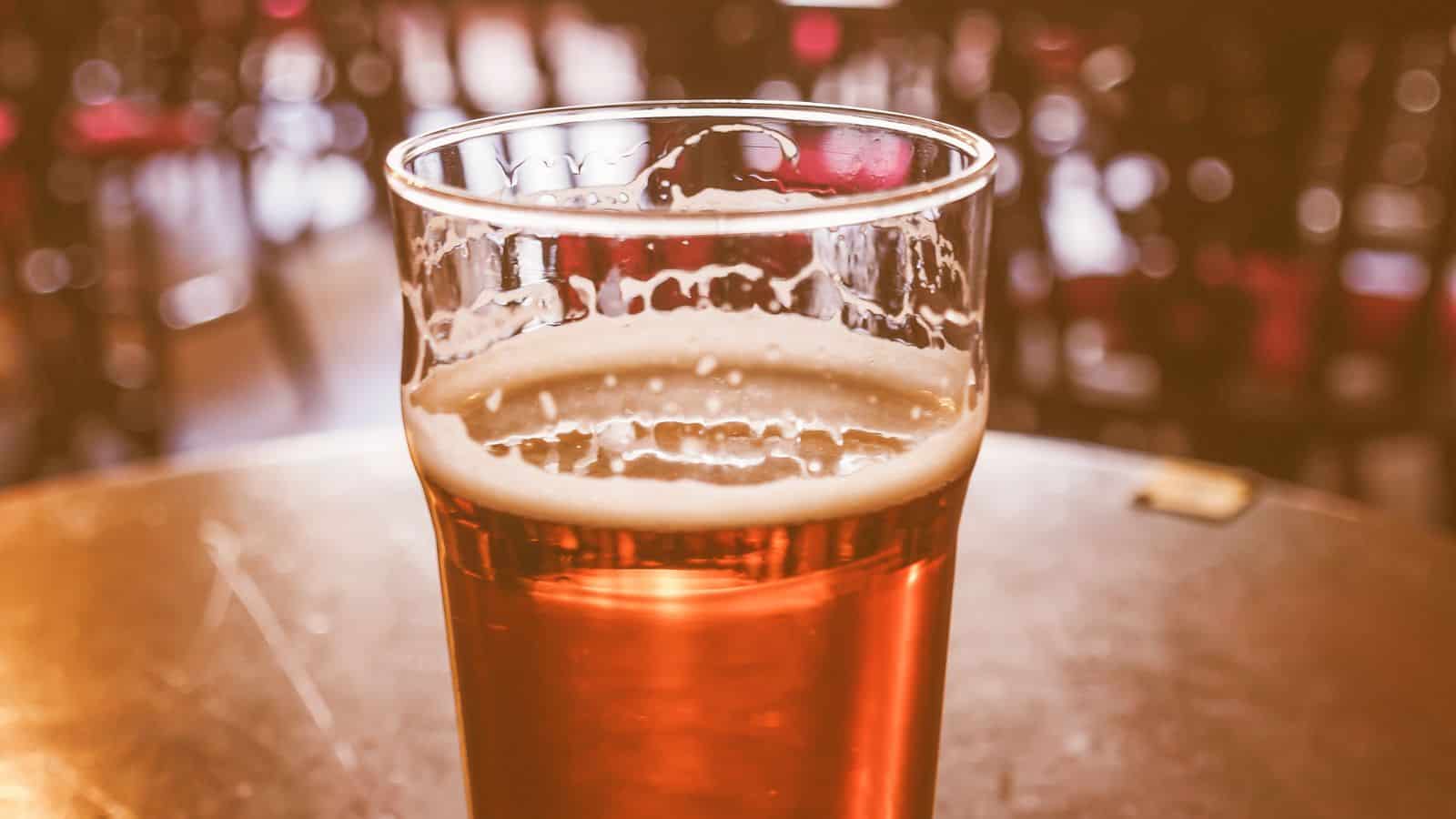 A close-up of a pint glass filled with amber beer, resting on a round table. The background is blurred, showing indistinct shapes of chairs and tables in a dimly lit setting.