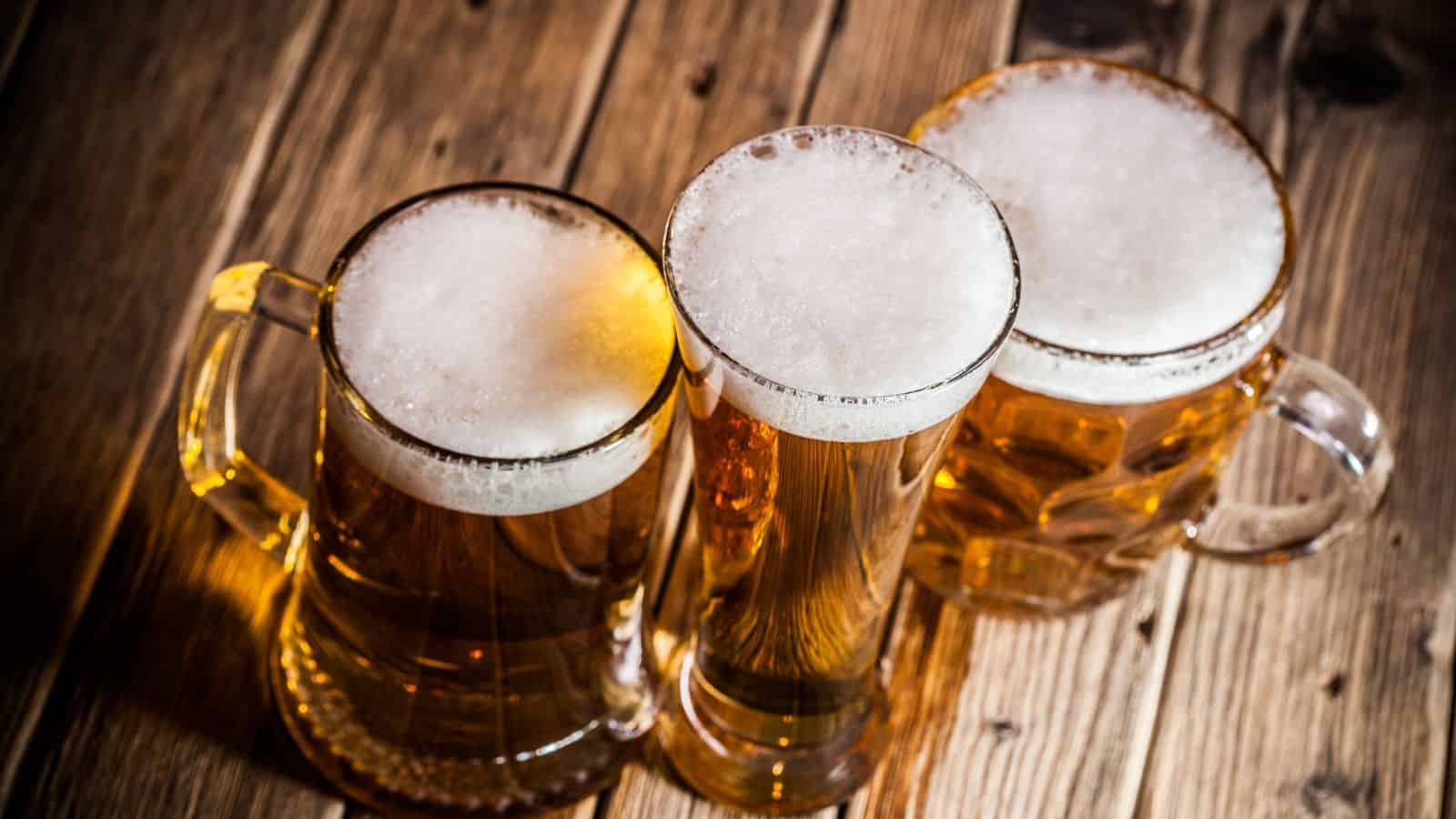 Three glass mugs of beer with frothy heads are placed closely together on a wooden surface, viewed from above. The mugs are filled with golden beer and the wood grain of the table is visible.