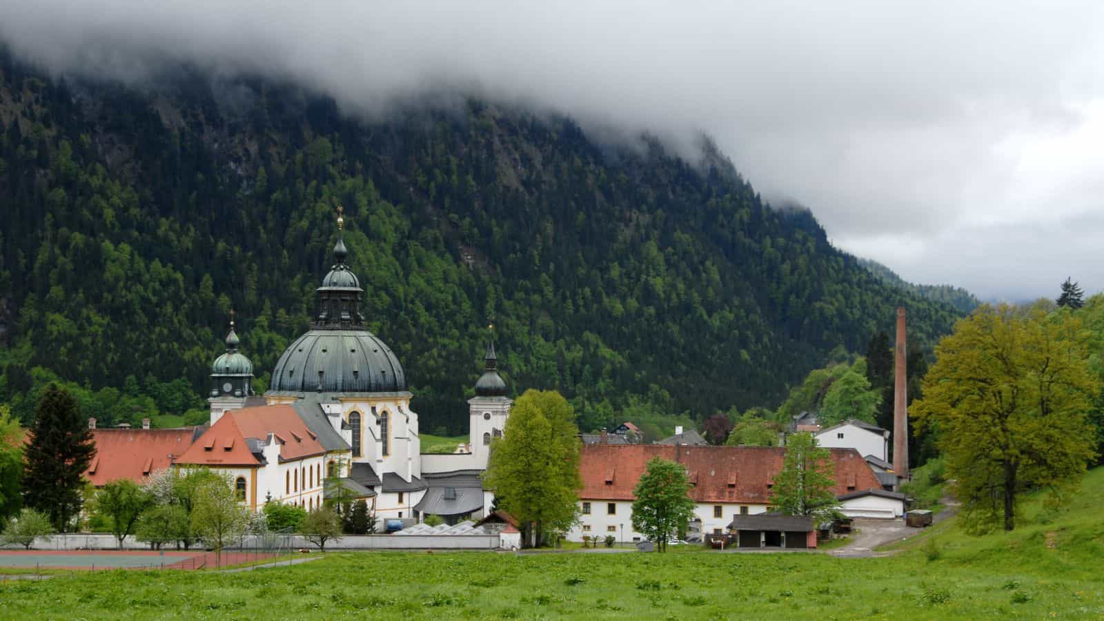 A historic monastery complex with a large domed church is surrounded by lush green trees and grass. It sits at the base of a forested mountain partially obscured by low clouds, creating a misty atmosphere.