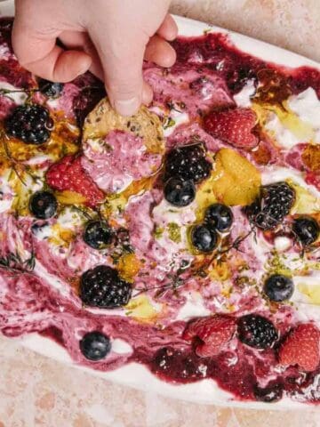 A hand dips a cracker into a platter of whipped cheese topped with a berry sauce, fresh berries, herbs, and olive oil, set on a beige marble surface. A plate with more crackers is visible in the corner.