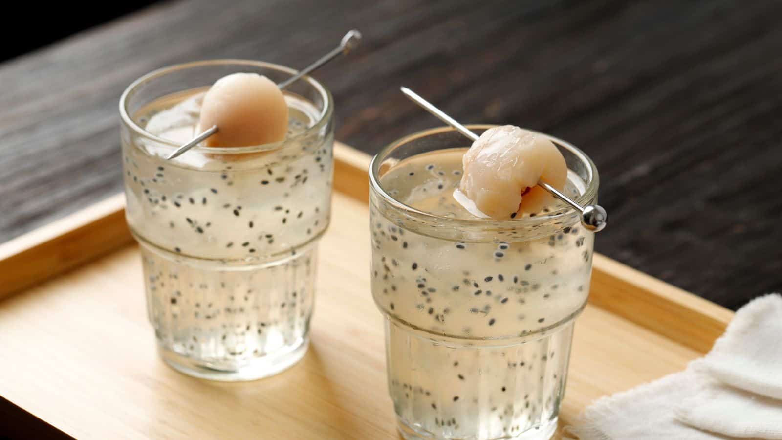 Two glasses of a clear beverage with chia seeds and ice cubes sit on a wooden tray. Each glass is garnished with a lychee on a metal skewer. A white cloth is on the tray's edge. The background is a dark wooden surface.