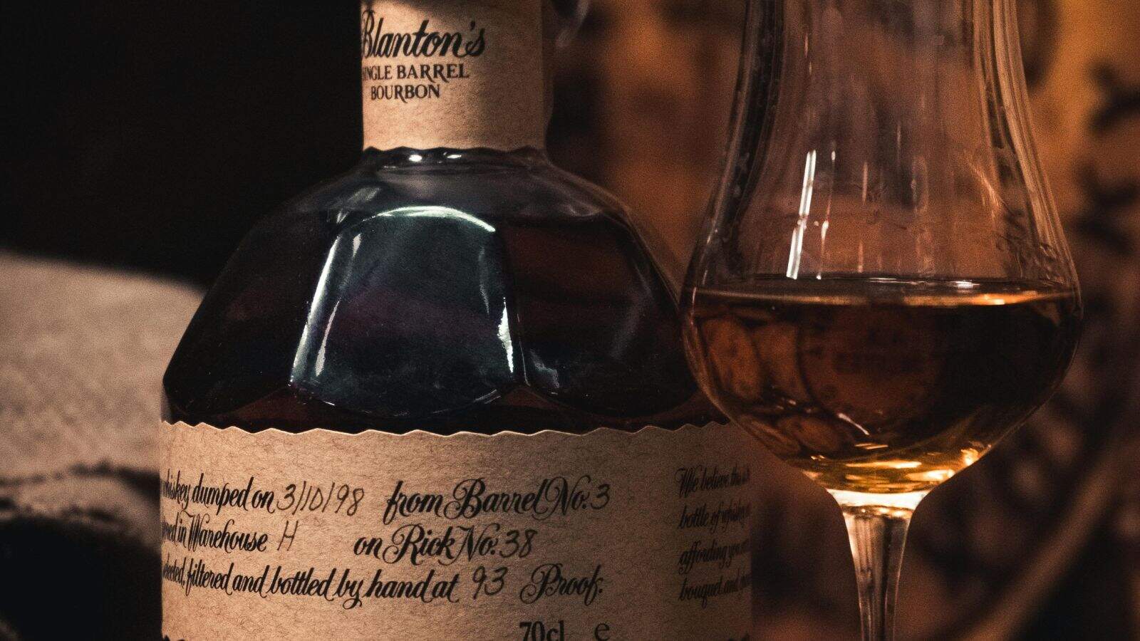 A close-up of a bottle of Blanton’s Single Barrel Bourbon with handwritten labeling next to a partially filled whiskey glass containing amber liquid. The background is softly blurred.