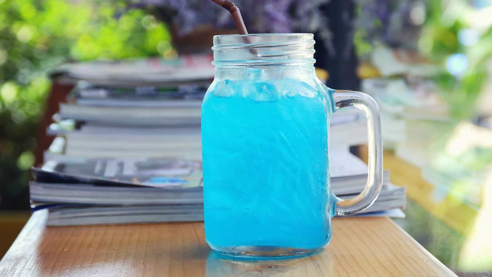 A mason jar filled with bright blue iced beverage and a brown straw sits on a wooden table, with a stack of magazines in the blurred background.
