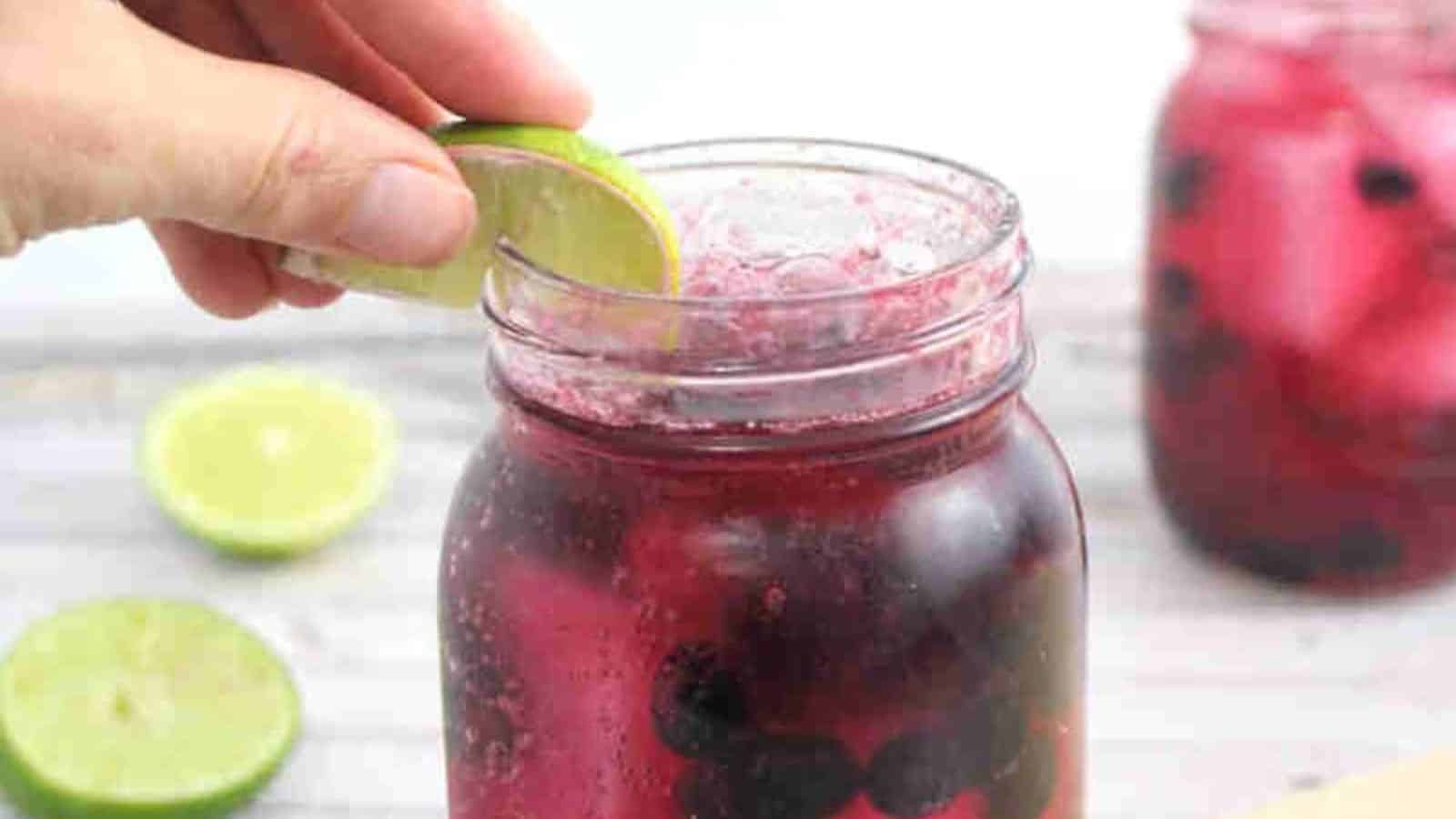 Blueberry mocktail in a mason jar with limes on the side of the jar and red straws with the jar on a wooden coater and cutting board and ingredients around it.