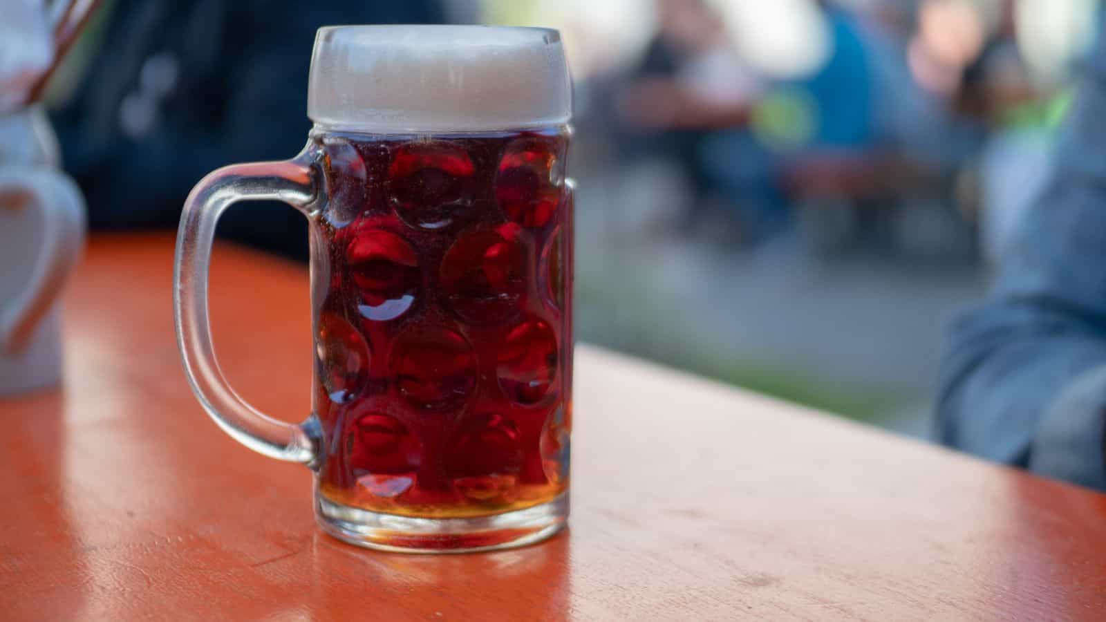 A glass mug filled with dark beer and topped with foam sits on an orange outdoor table, with blurred people and seating visible in the background.