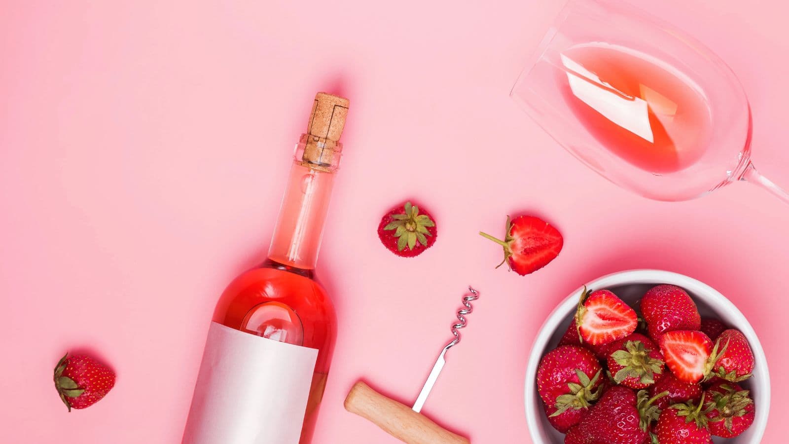 A bottle of rosé wine with a cork, a corkscrew, a glass of rosé, a bowl of strawberries, and loose strawberries are arranged on a pink surface.
