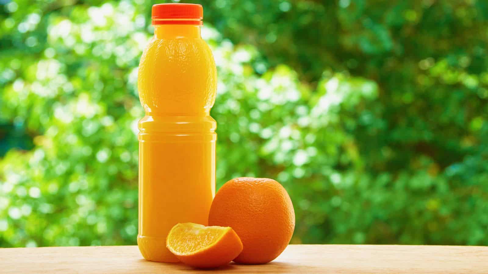 A plastic bottle of orange juice with an orange cap stands on a wooden surface next to a whole orange and a wedge of orange. Green foliage is blurred in the background.