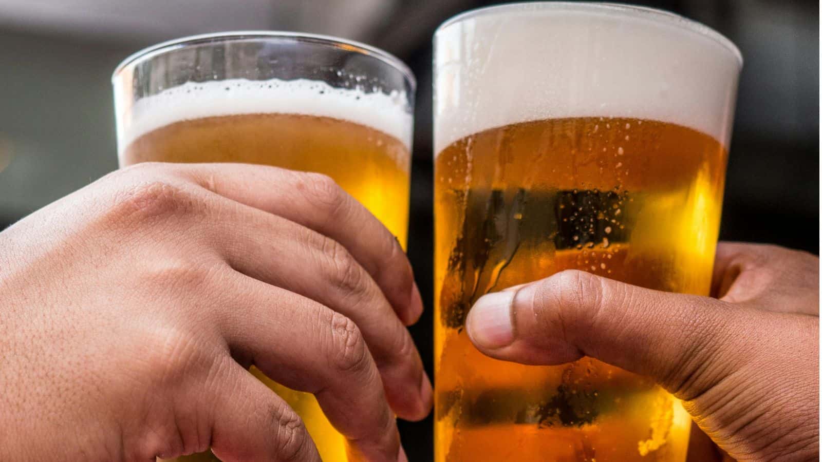 Two hands holding pint glasses filled with amber-colored beer are clinking together. The beer has a frothy white head. The background is blurred, focusing attention on the glasses.