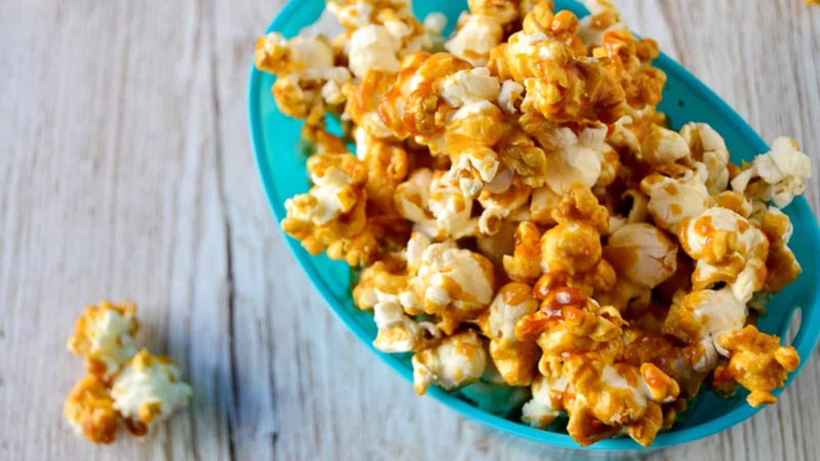 A teal bowl filled with caramel-coated popcorn is placed on a wooden surface. A few pieces of popcorn are scattered beside the bowl.