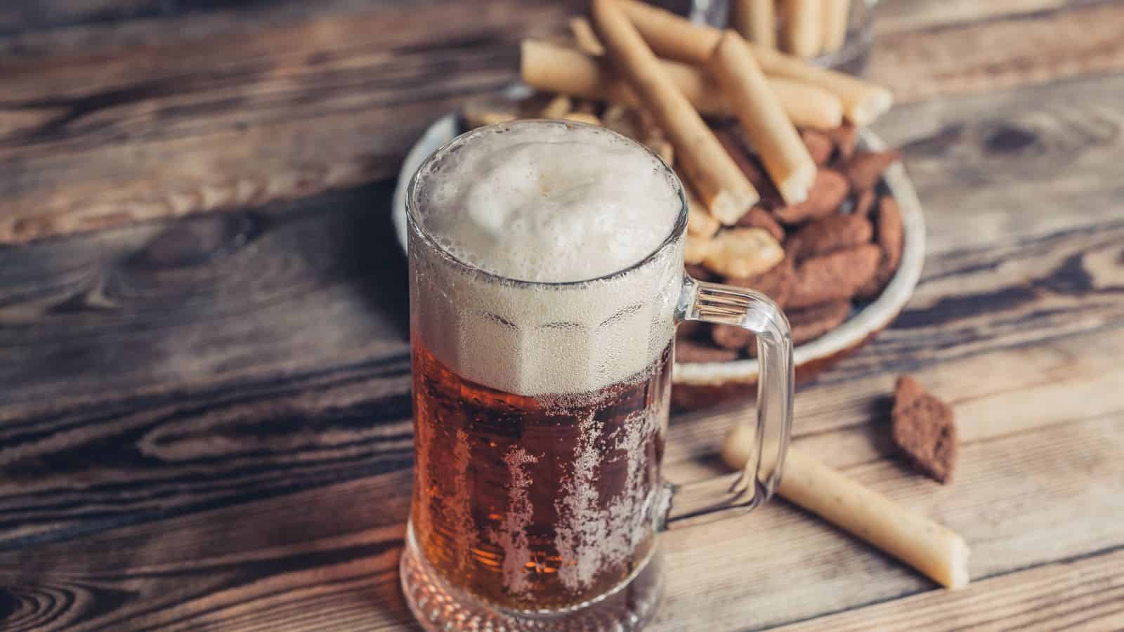A mug of frothy beer on a wooden table, with a plate of breadsticks and pretzels in the background.
