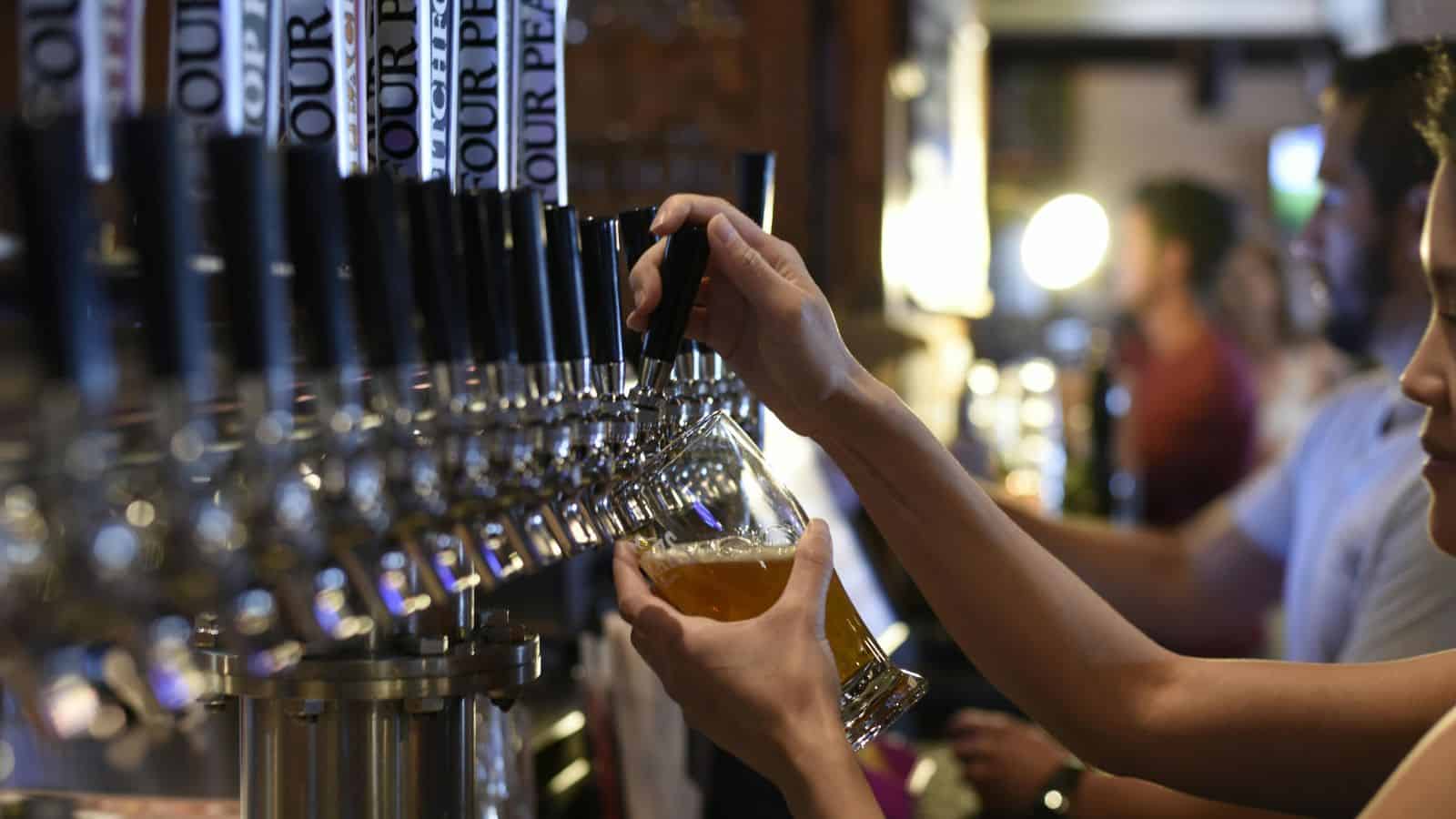 A person pours beer from one of many taps into a glass at a bar. Several taps are lined up in a row, and other people are visible in the background.