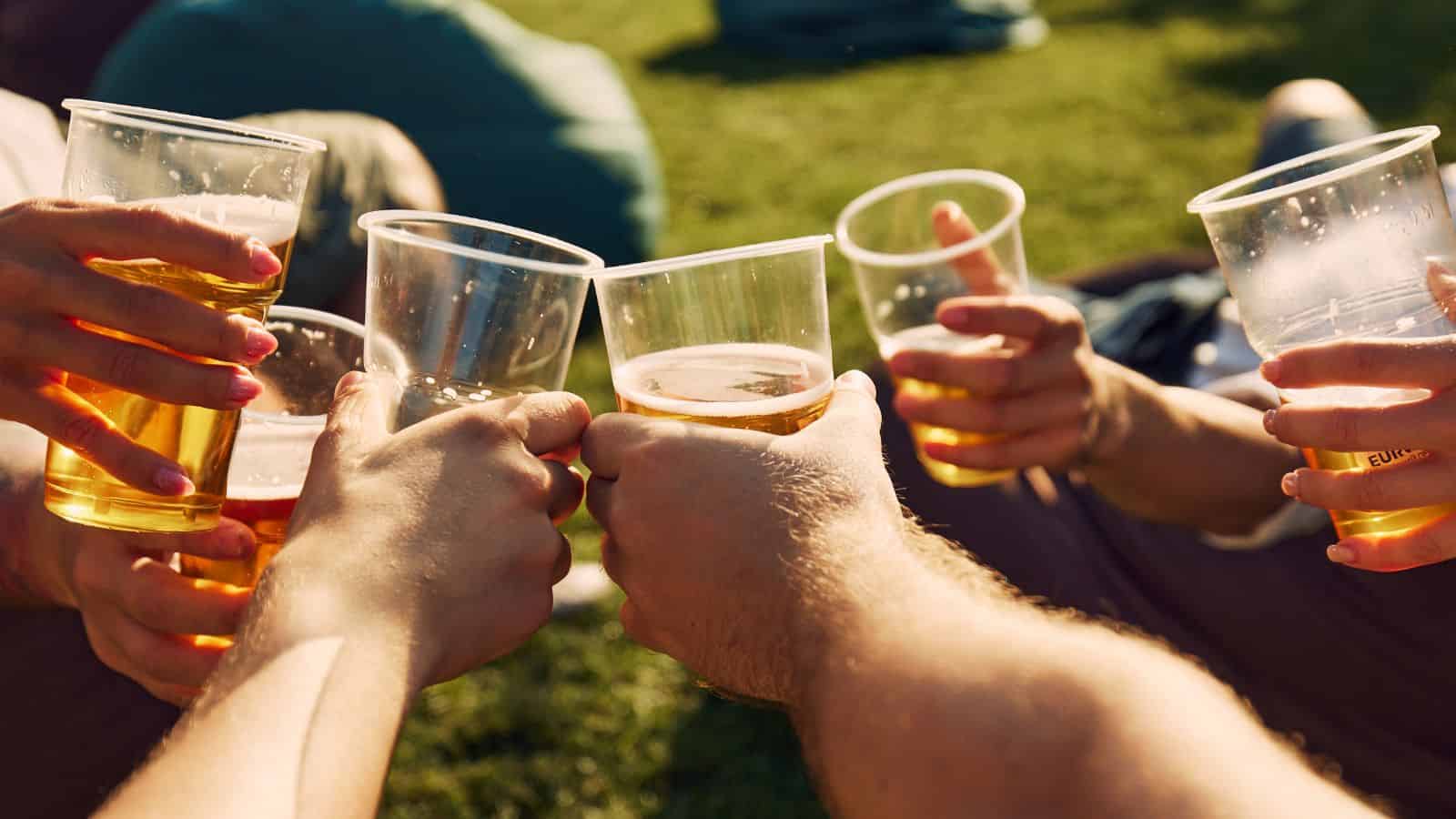Several hands holding plastic cups filled with beer clink their drinks together outdoors on a sunny day, with green grass visible in the background.