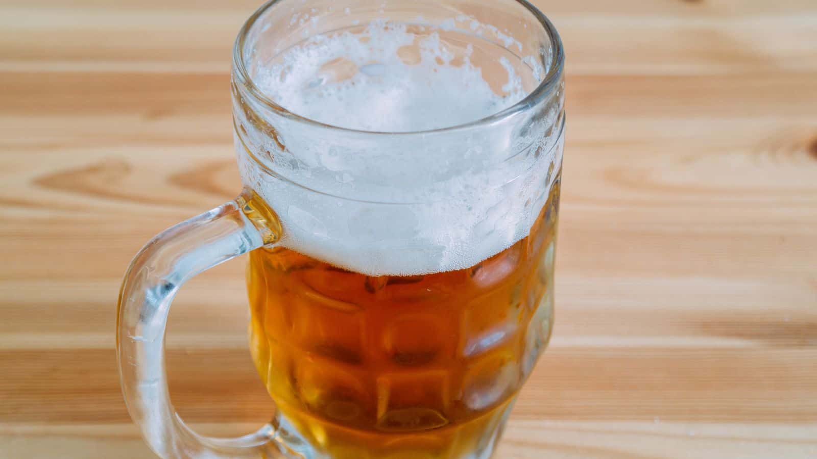 A glass mug filled with light amber beer topped with a layer of foam, placed on a wooden surface.