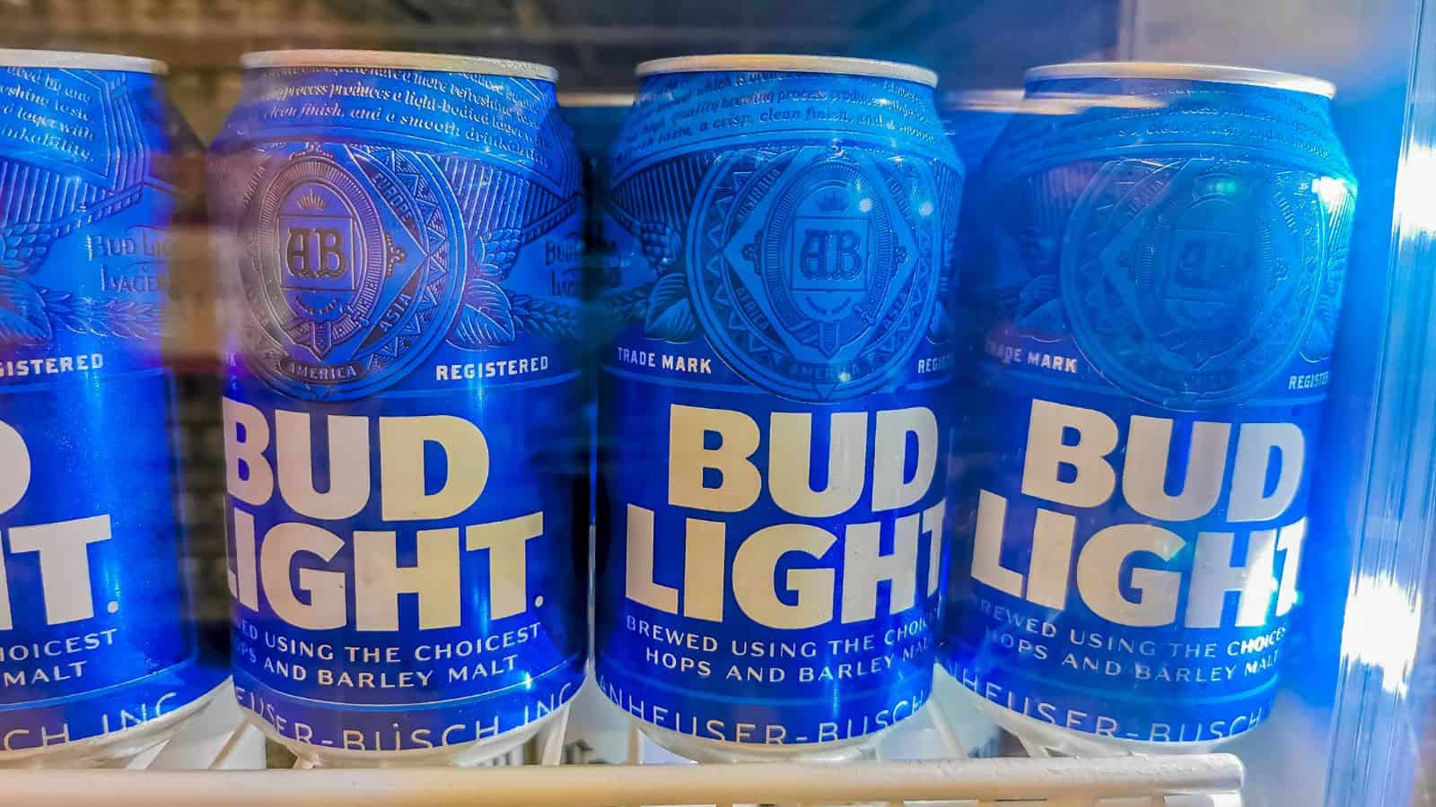 Several blue Bud Light beer cans are arranged in a refrigerator. The cans feature the Bud Light logo in large white letters and display details about the beer’s ingredients and branding.