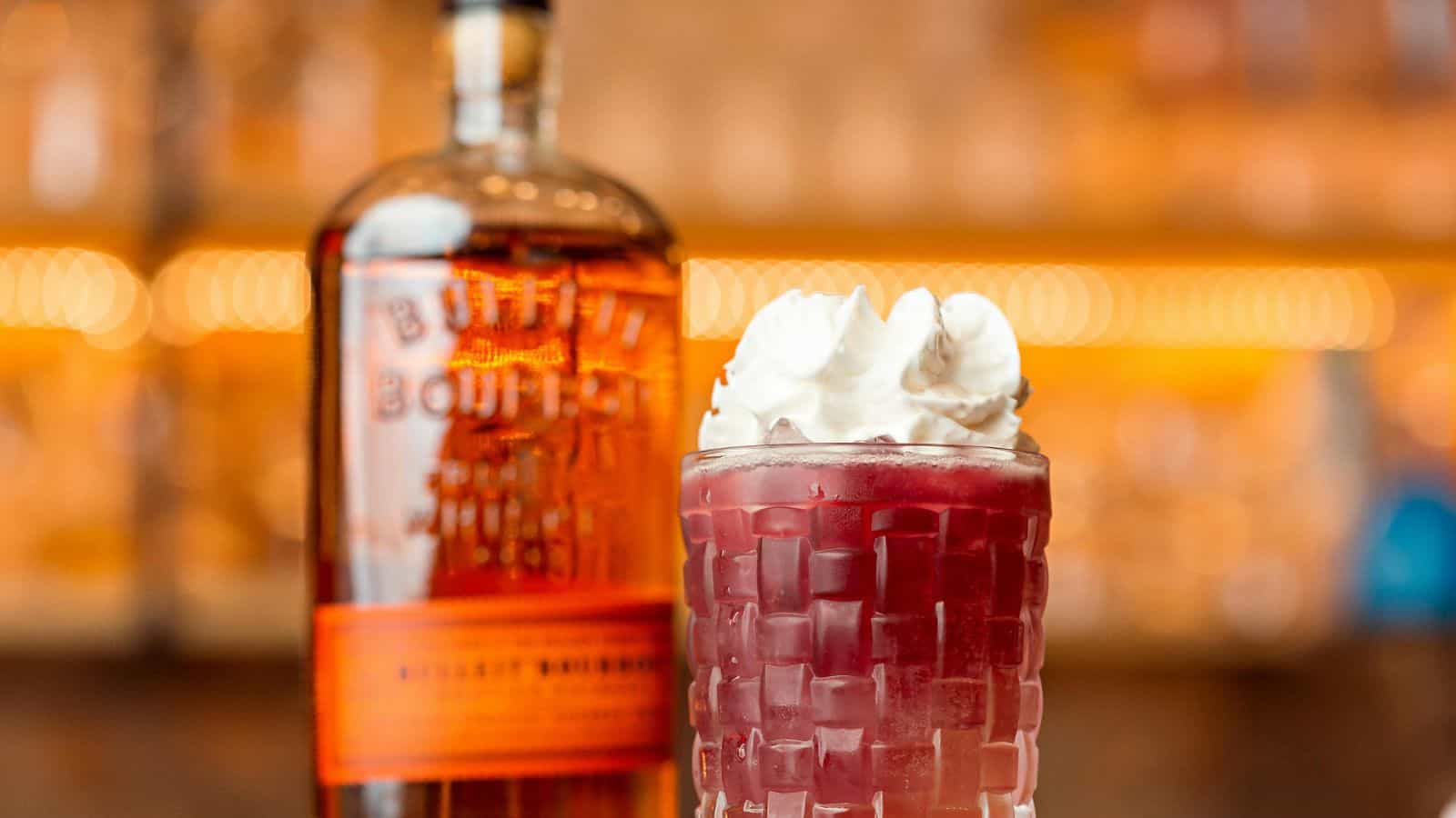 A glass filled with a dark red beverage topped with whipped cream sits in front of a blurred bottle of bourbon whiskey on a bar counter.
