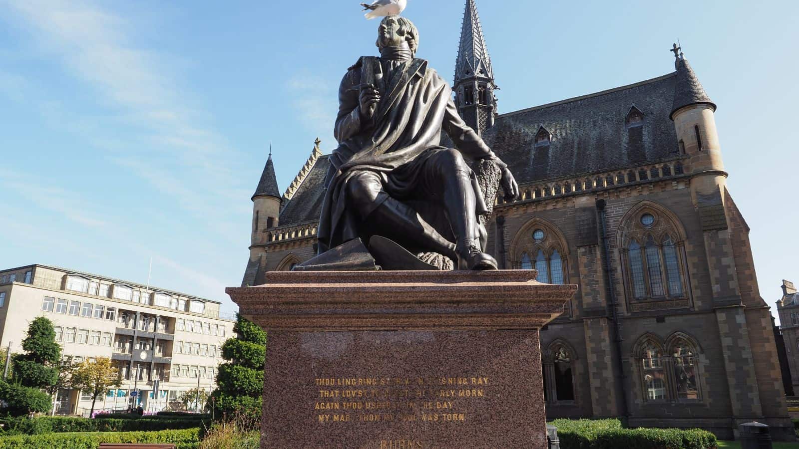 A bronze statue of a seated man on a stone pedestal stands in front of a large church-like building with arched windows and spires. A white bird perches on the statue&rsquo;s head. Trees and other buildings are in the background.