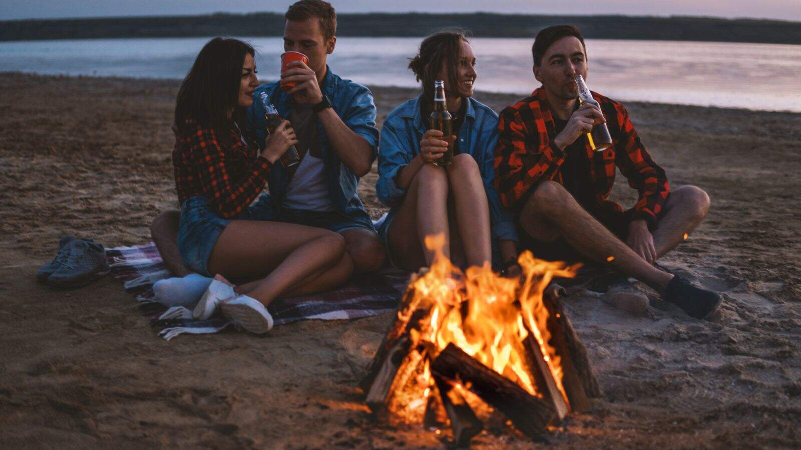 Four people sit on a blanket around a campfire on a sandy beach at sunset, drinking from bottles and cups. The group appears relaxed, with a lake and distant shoreline in the background.