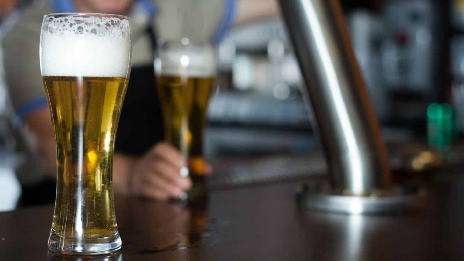A close-up of two frosty glasses filled with light beer on a bar counter. The bar has a shiny metal draft tower, and there are blurred figures in the background, suggesting a lively atmosphere.