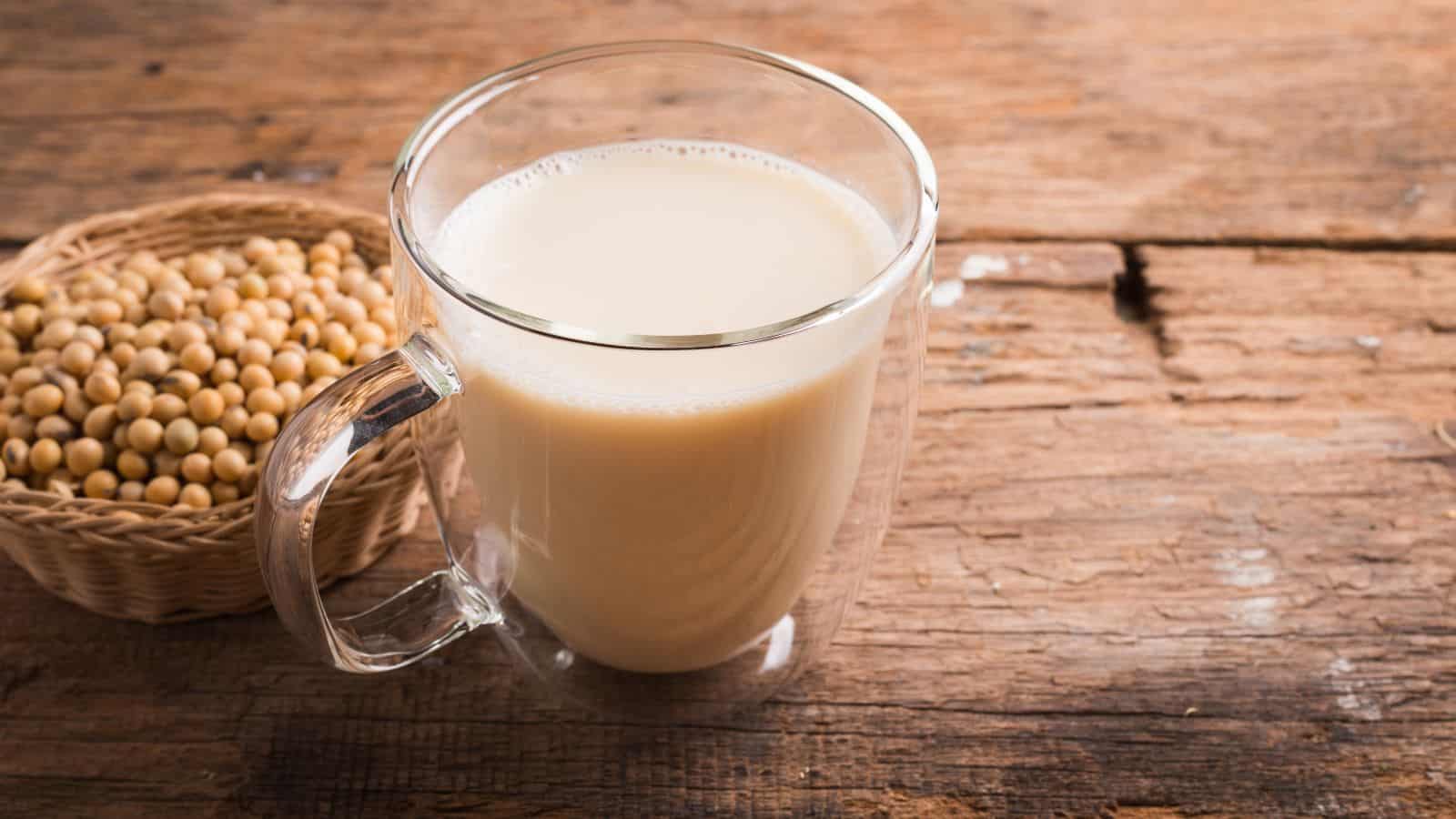 A glass cup of soy milk is placed on a wooden surface. Next to it is a small woven basket filled with soybeans. The wooden background has a rustic and textured appearance.