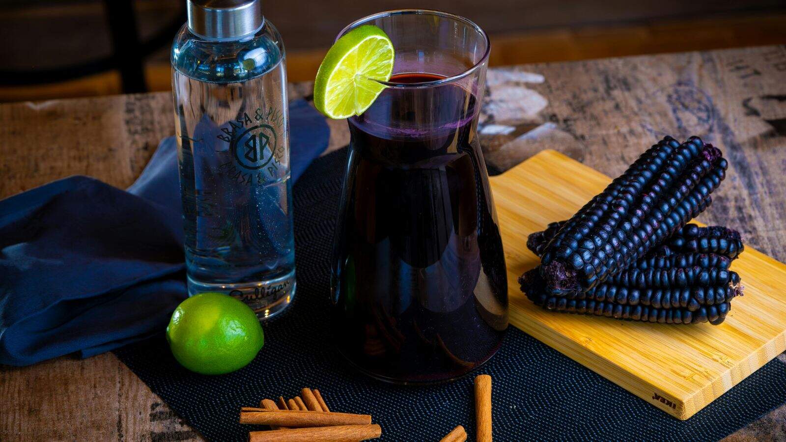 A wooden table with a carafe of purple liquid garnished with a lime slice, a bottle of clear liquid, a lime, a bundle of purple corn on a cutting board, cinnamon sticks, and a dark napkin.