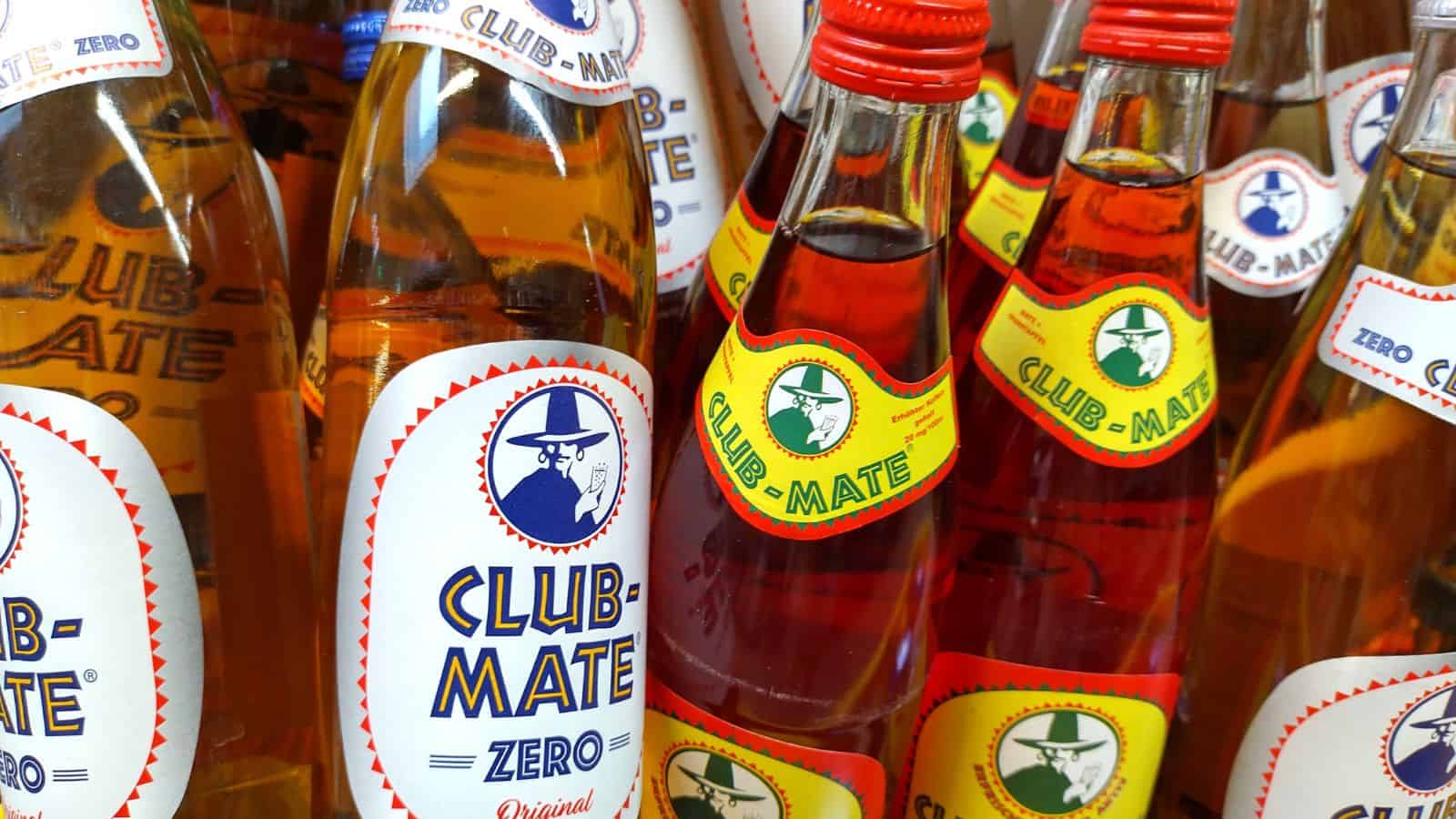 Bottles of Club-Mate, a carbonated beverage, are displayed closely together. The bottles feature red and blue caps, with labels in white and yellow showing a logo of a person wearing a hat. Some bottles are labeled "Zero.