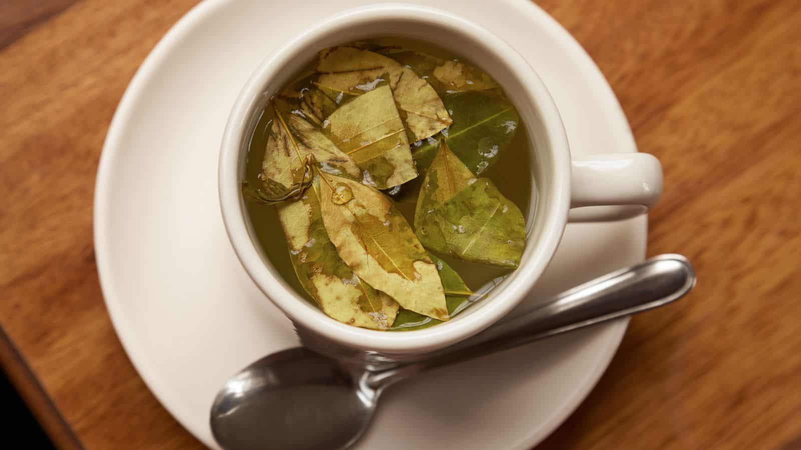 A cup filled with a hot beverage containing steeped leaves is placed on a white saucer. A metal spoon rests on the saucer beside the cup. The scene is set on a wooden table.