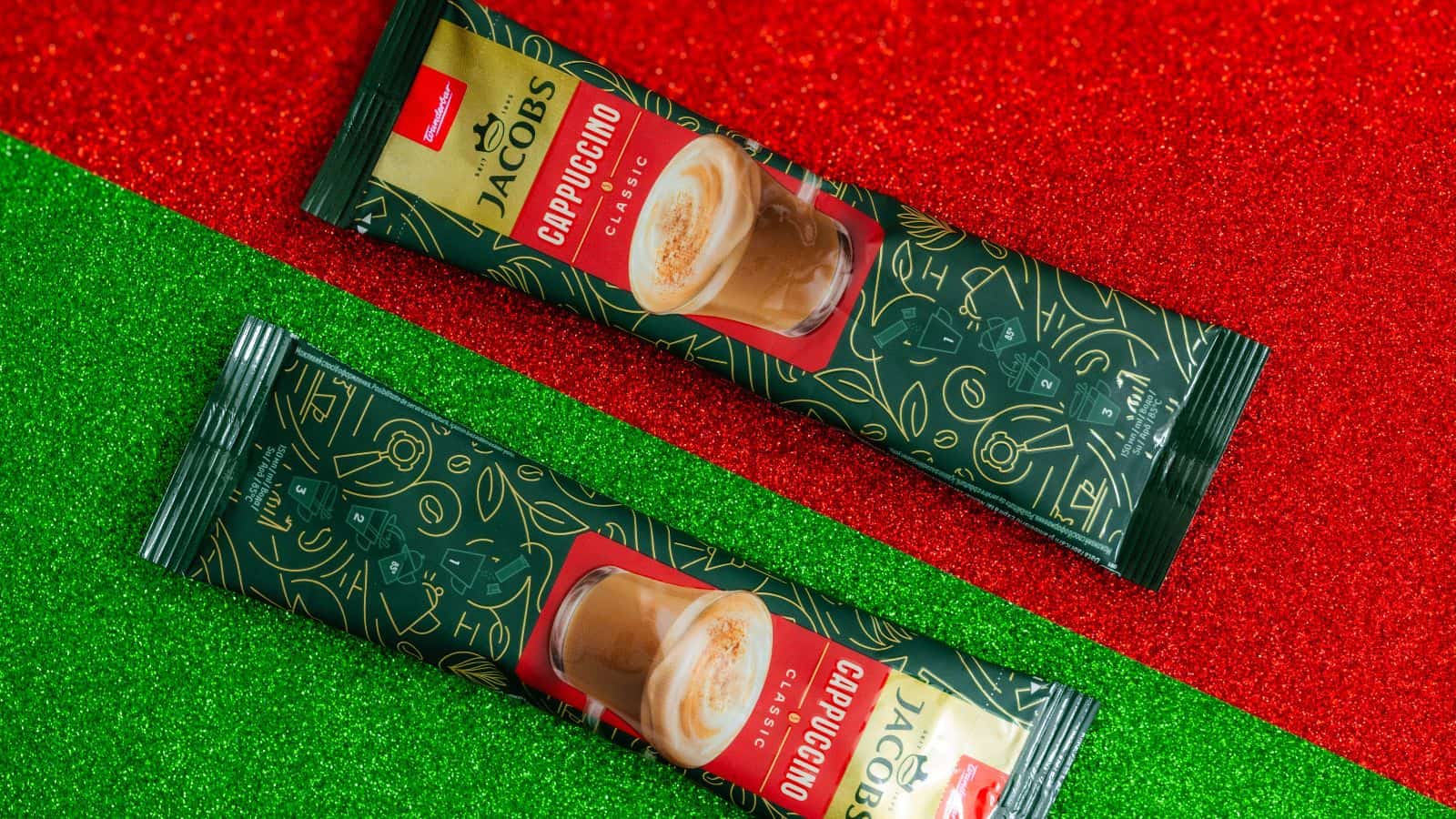 Two packets of Jacobs Cappuccino Original instant coffee are placed diagonally on a glittery background divided into red and green sections. The packaging is dark with gold designs and features an image of a cappuccino cup.