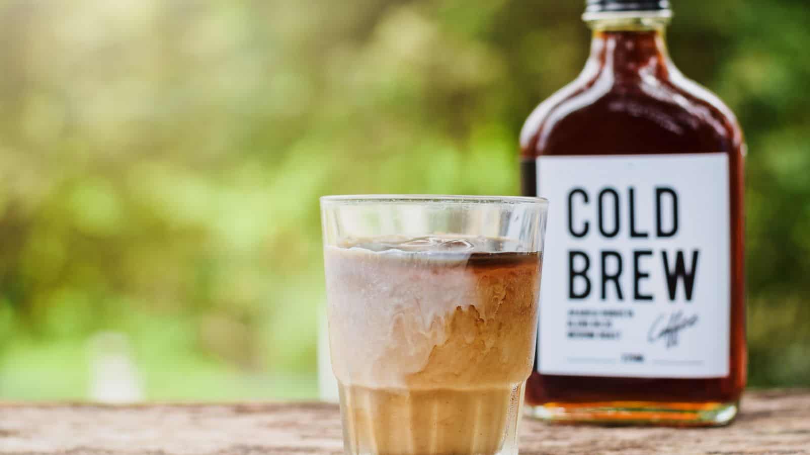 A glass of iced cold brew coffee with milk sits on a wooden surface. In the background, there is a bottle labeled "Cold Brew." The setting appears to be outdoors with greenery visible.