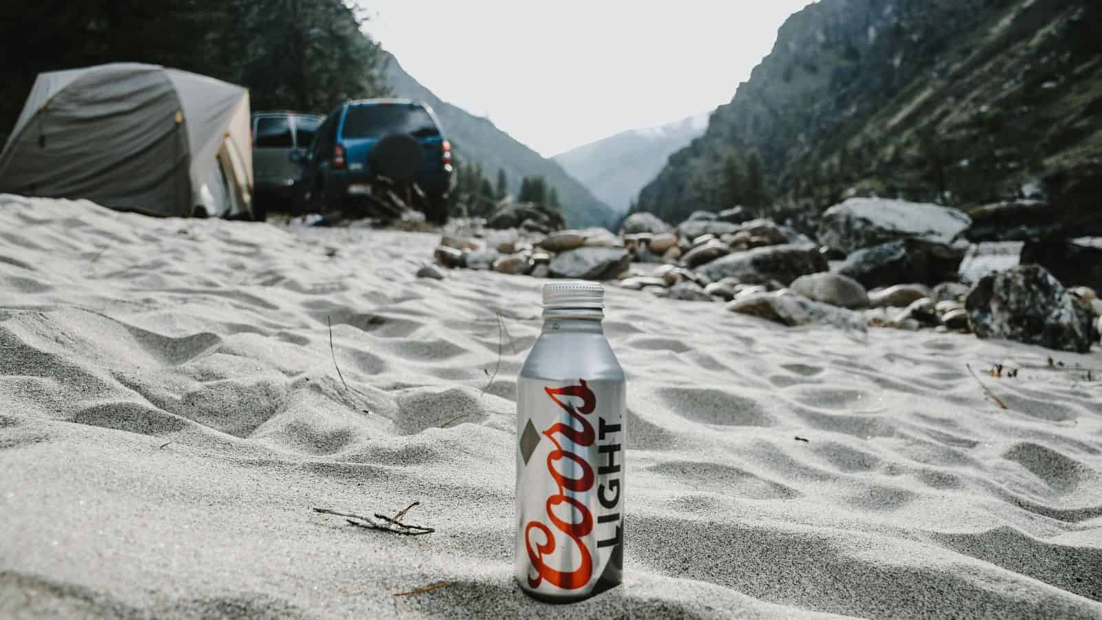 A Coors Light aluminum bottle stands upright on sandy ground at a campsite, with a tent, vehicles, rocks, and mountains in the background under a partly cloudy sky.