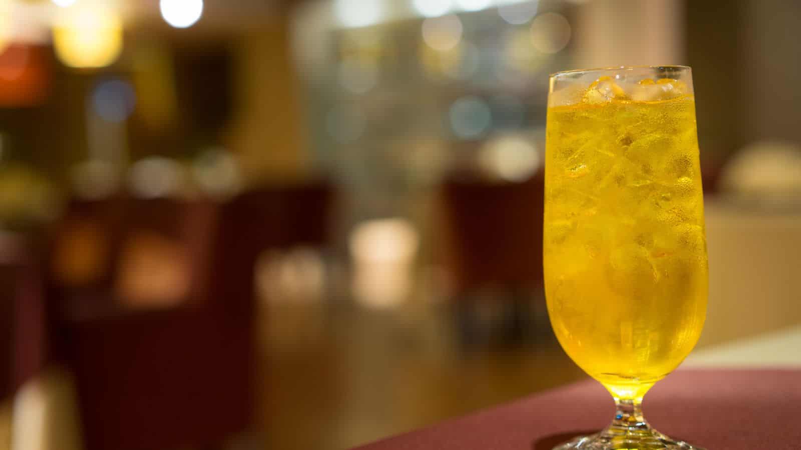 A tall glass filled with an orange-yellow iced beverage sits on a table. Ice cubes are visible in the drink. The background is softly blurred, showing a warmly lit indoor setting with tables and chairs.