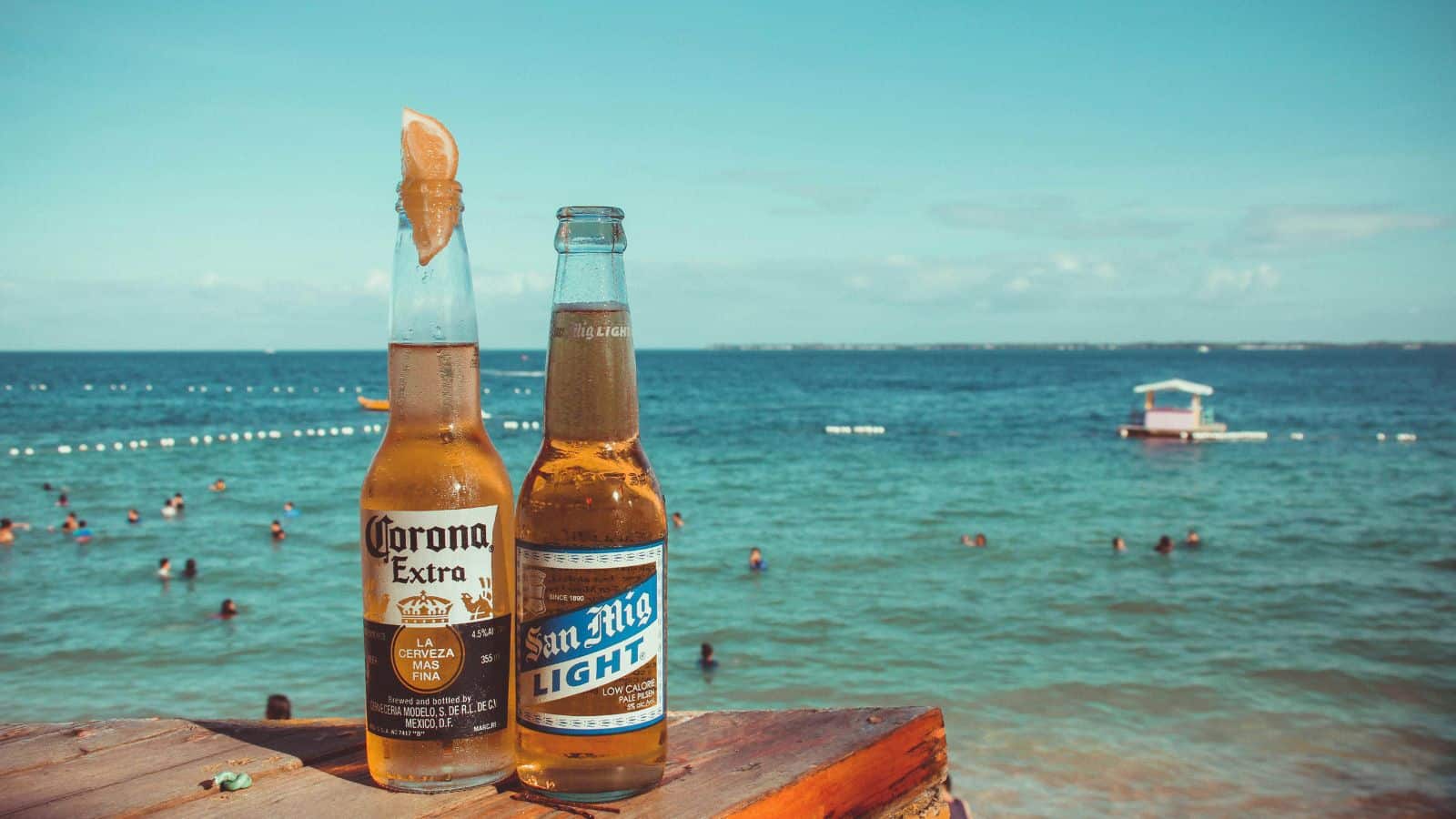 Two bottles of beer, one Corona Extra with a lime wedge and one San Mig Light, are on a wooden surface by the beach. People are swimming in the sea, and a small boat is visible in the background under a clear sky.