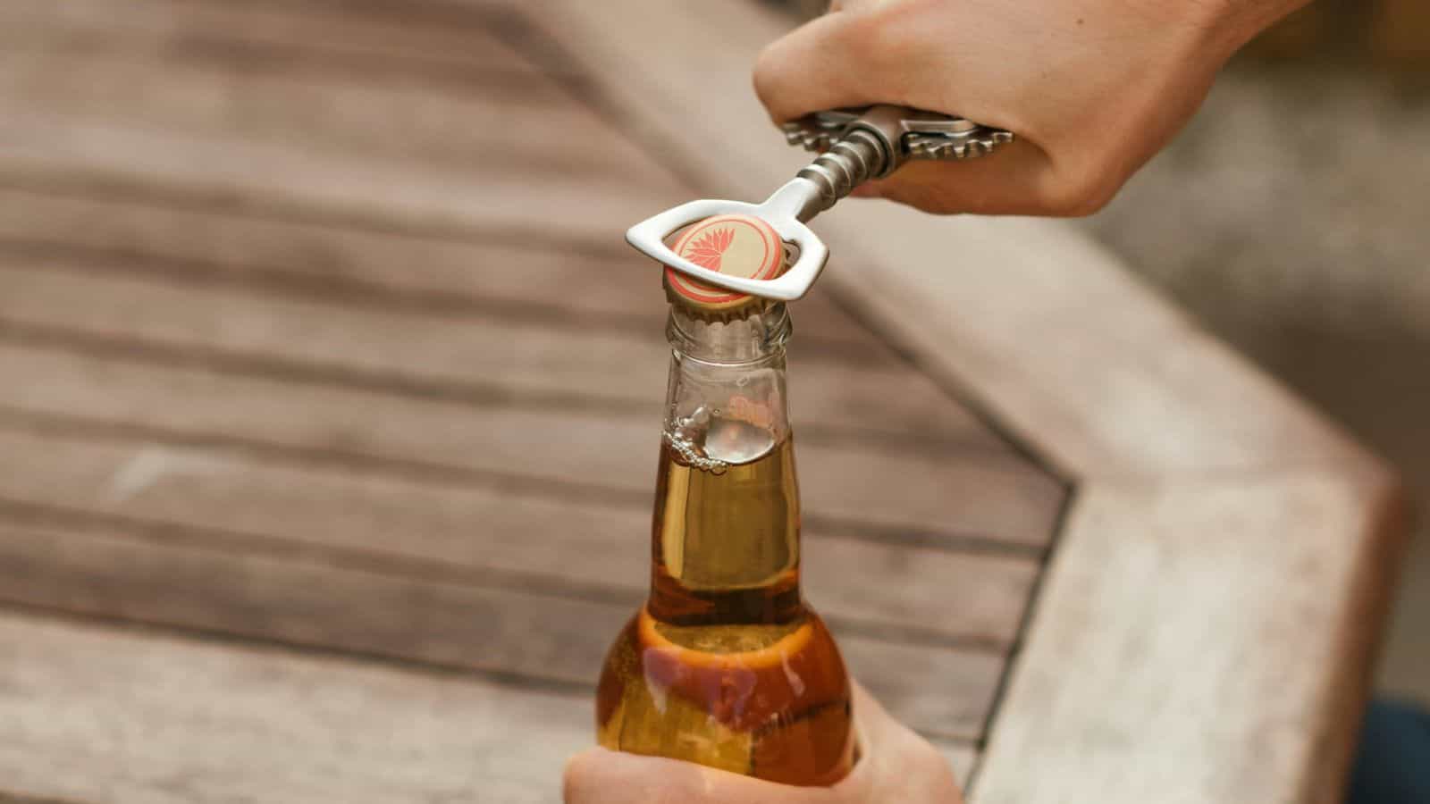 A person uses a metal bottle opener to open a capped glass bottle of beer on a wooden table.