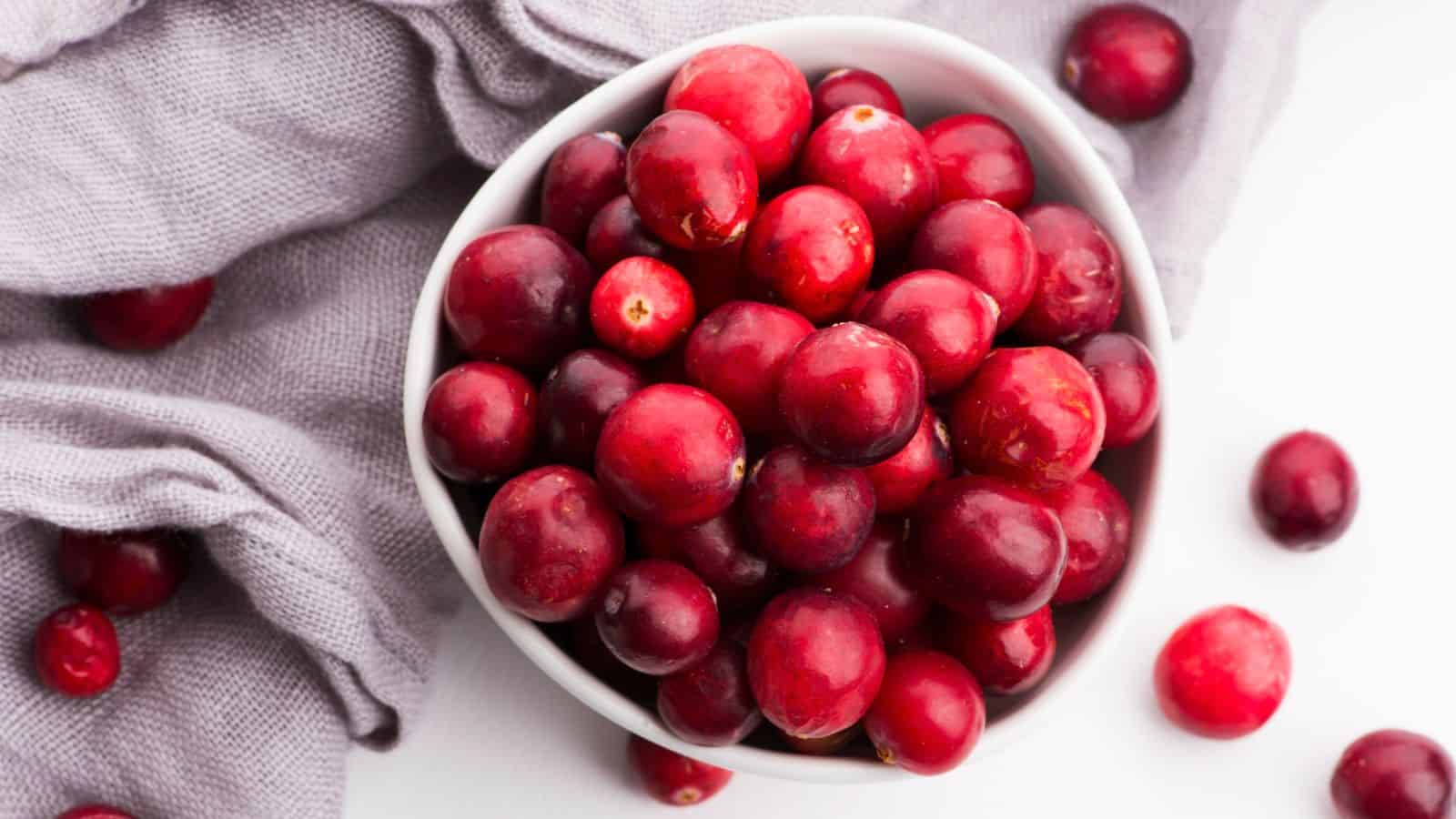 A white bowl filled with fresh red cranberries sits on a white surface next to a light gray cloth, with a few loose cranberries scattered nearby.