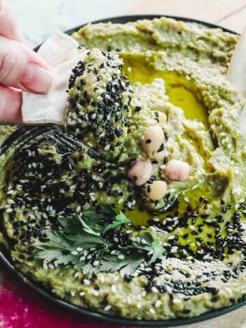 A bowl of green hummus garnished with black sesame seeds, chickpeas, and herbs sits on a wooden board. A hand dips a piece of pita bread into the hummus. Slices of pink and white radish are visible on the board.
