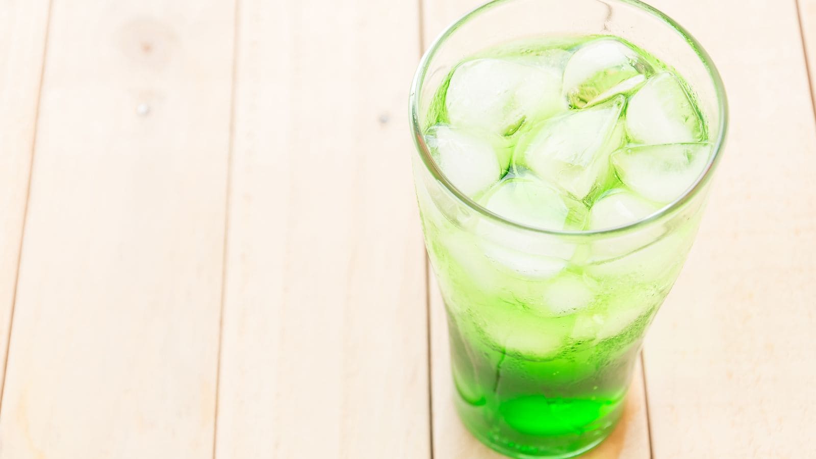 A glass filled with green liquid and ice cubes is on a light wooden surface. The drink is bubbly, and the ice creates a refreshing appearance.