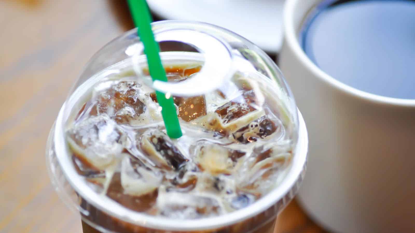 Close-up of a plastic cup with iced coffee and a green straw. The cup has a plastic lid and is filled with ice cubes. In the background, there is a white ceramic cup filled with dark coffee on a wooden surface.