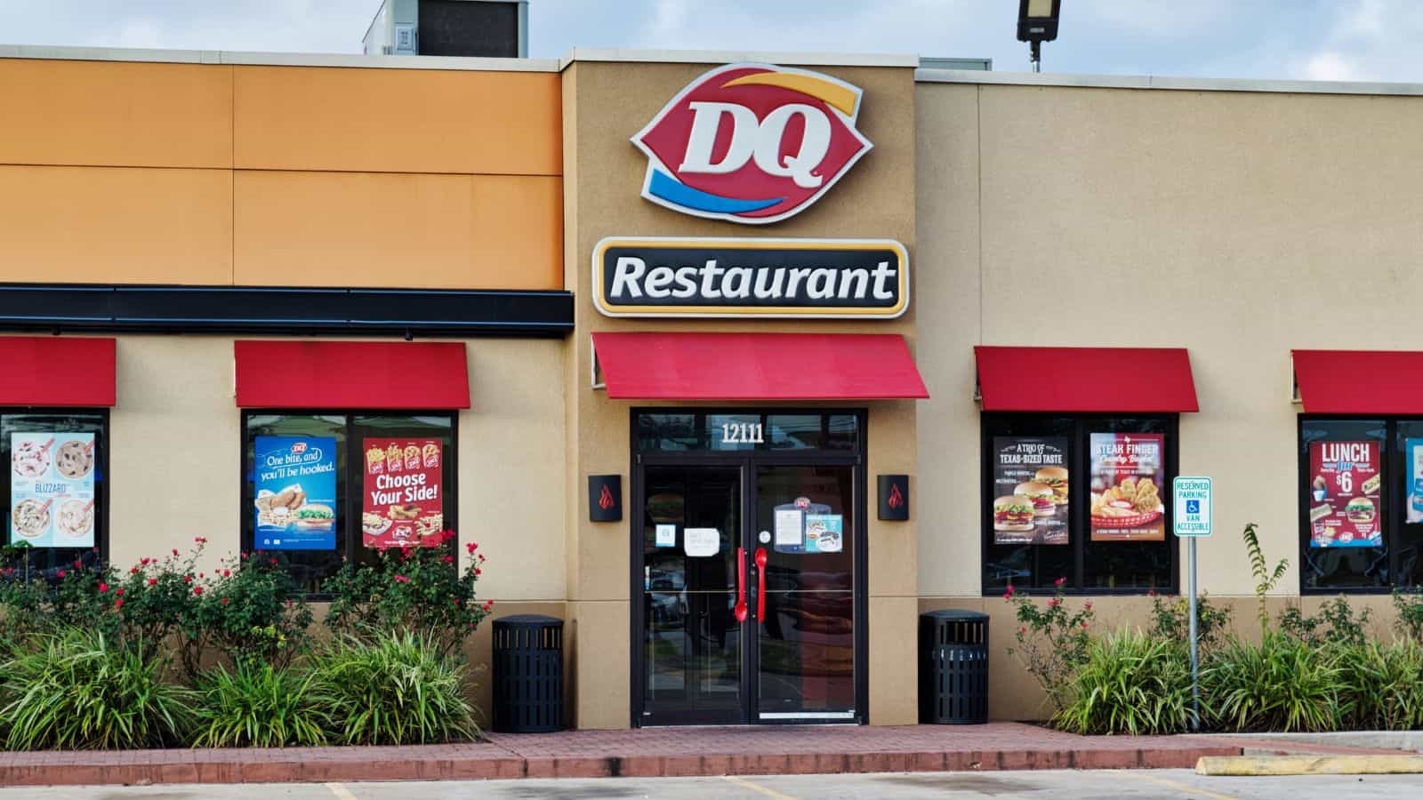 The front entrance of a Dairy Queen restaurant with red awnings, several menu posters on the windows, plants along the building, and a sign above the door displaying the “DQ Restaurant” logo.