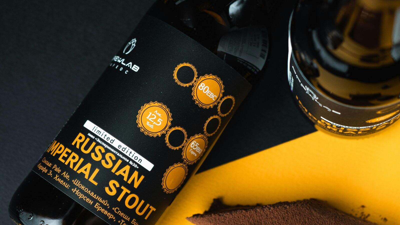 Close-up of two bottles of limited edition Russian Imperial Stout on a black and yellow background. The label shows measurements like 12.5% ABV and 65 IBU. A slice of chocolate cake is partially visible at the bottom.