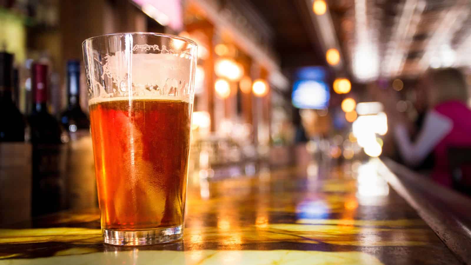 A pint glass filled with amber-colored beer sits on a polished bar counter in a dimly lit bar, with blurred bottles and patrons visible in the background.