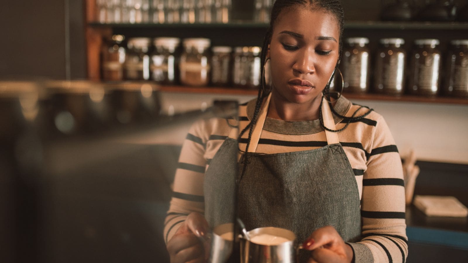 A person wearing a striped shirt and apron is holding a milk frothing pitcher, looking down at it intently. They are standing in a cafe setting with shelves of jars in the background.