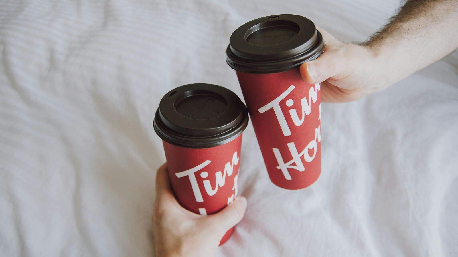 Two hands are holding red Tim Hortons coffee cups with brown plastic lids. The background shows a white, textured surface, likely a bedspread or tablecloth. Both cups display the brand's logo in white text.
