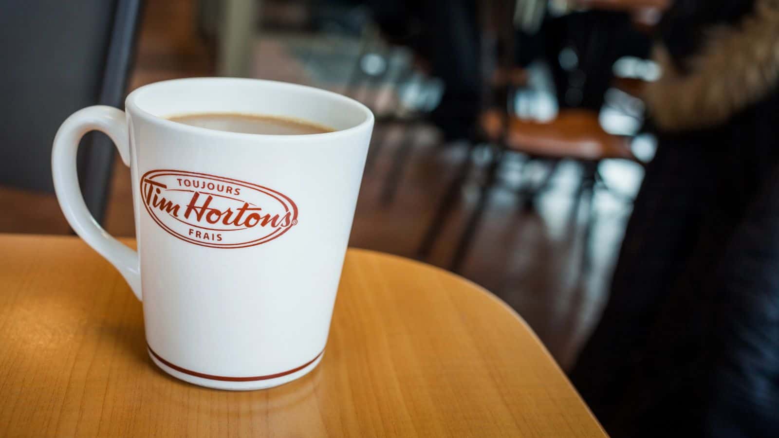 A white Tim Hortons mug filled with coffee sits on a wooden table. The mug features the Tim Hortons logo in red. The background shows a blurred indoor cafe setting with wooden chairs and a person's coat.