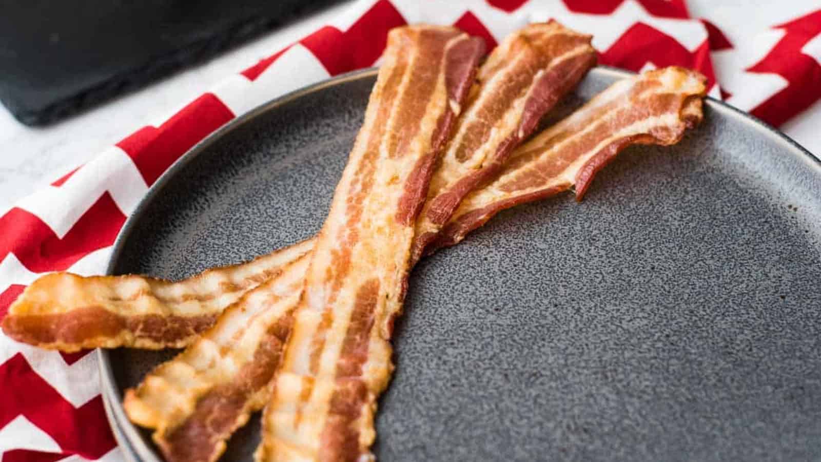 Four strips of crispy bacon are neatly arranged on a gray plate. The plate rests on a red and white zigzag-patterned napkin. In the background, there is a black cooling rack with a wooden spatula on it.
