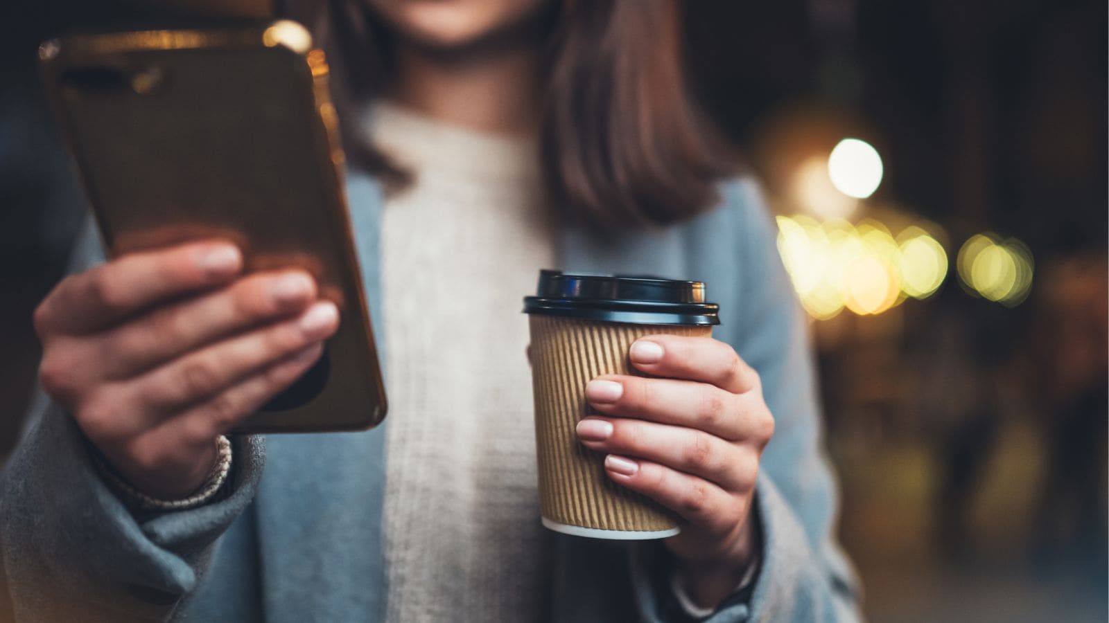A person wearing a gray coat holds a smartphone in one hand and a disposable coffee cup in the other. The background is blurred with warm lights, suggesting an outdoor or evening setting.