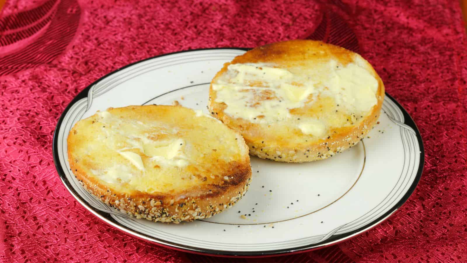 A plate holds two halves of a toasted bagel with butter spread on top. The bagel has a sesame and poppy seed coating and rests on a white plate with a patterned edge. The background is a textured red surface.