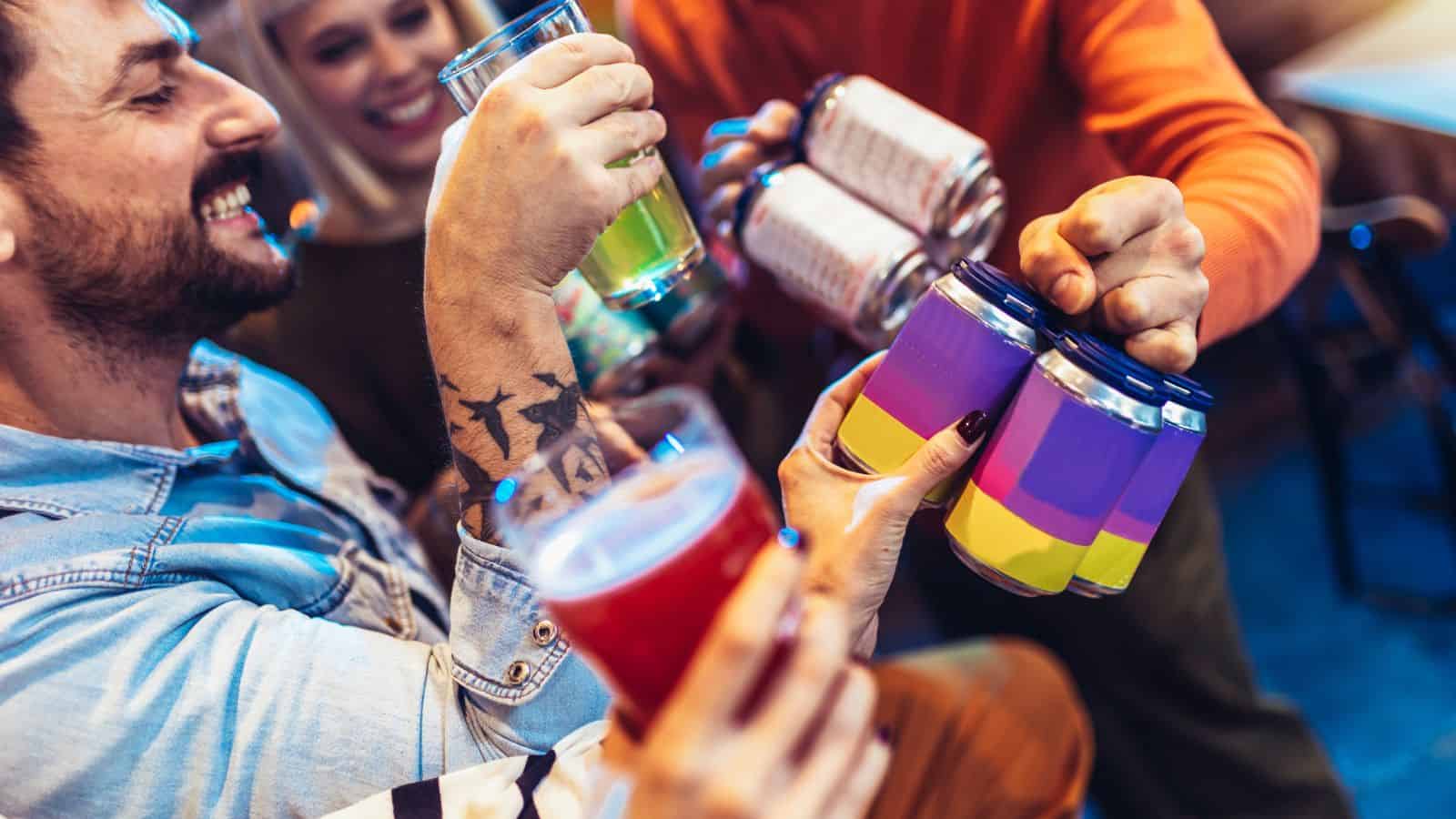 A group of people clink glasses and cans together in a toast. The drinks include colorful beverages in clear glasses and canned drinks with purple and yellow labels. The scene is lively and social.