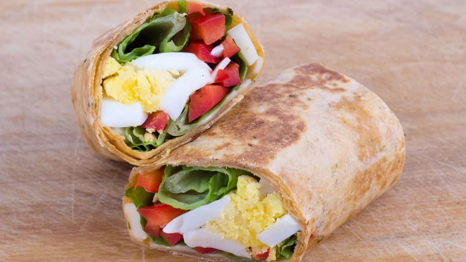 A wrap cut in half, displayed on a wooden surface. It contains sliced boiled eggs, lettuce, red bell peppers, and cheese, all wrapped in a toasted flatbread.