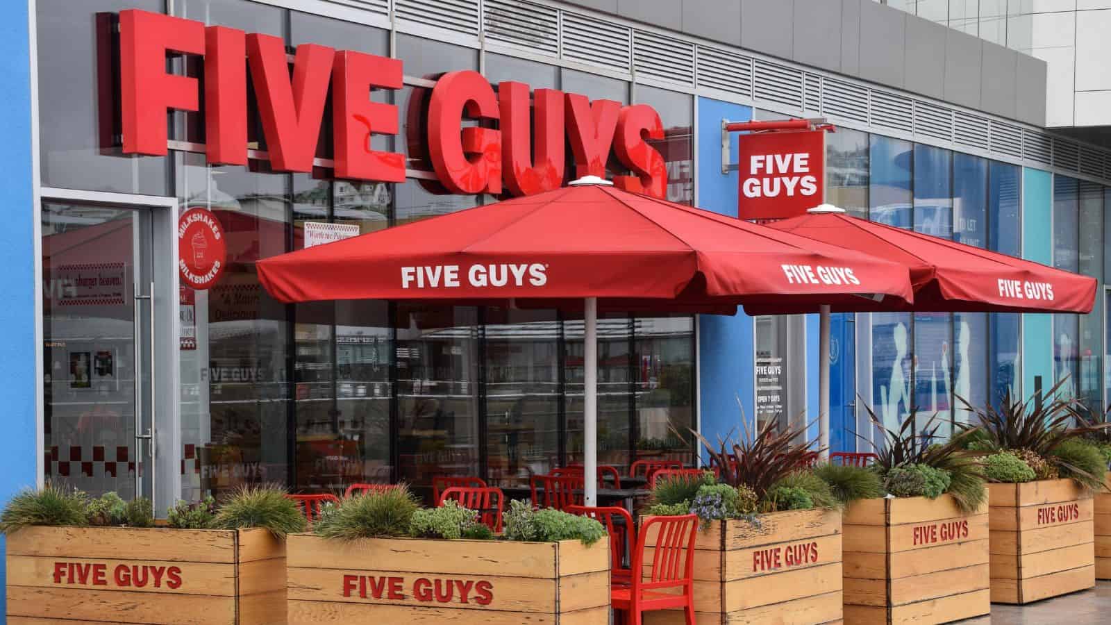 The exterior of a Five Guys restaurant with large red signage, red umbrellas, red chairs, and wooden planters in front of the outdoor seating area. The storefront has large glass windows.